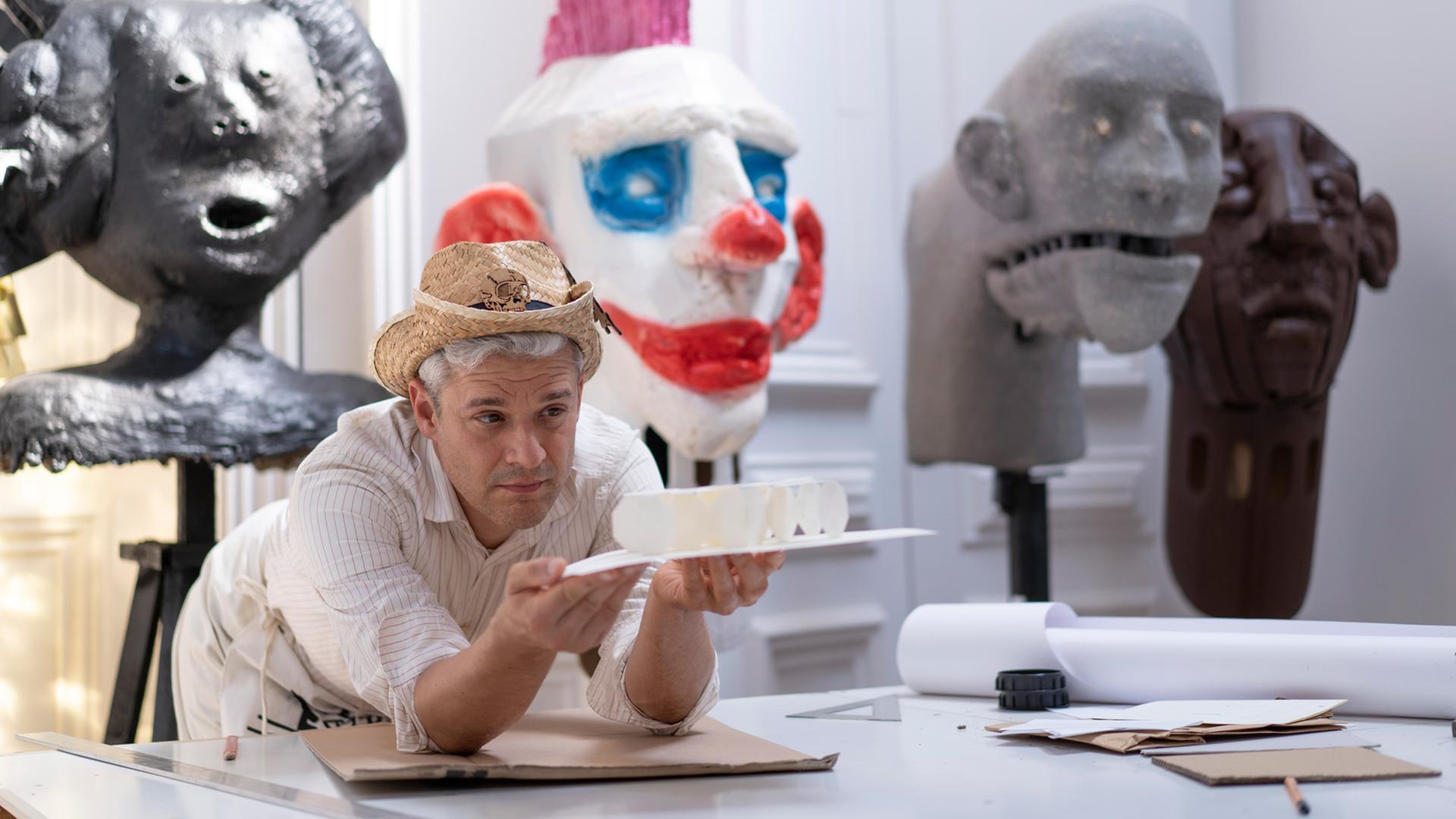 Tobias Binderberger stützt sich in seinem Atelier auf einen Tisch und hält ein Kunstobjekt in der Hand. Auf dem Kopf trägt er einen Strohhut. Tobias Binderberger stützt sich in seinem Atelier auf einen Tisch und hält ein Kunstobjekt in der Hand. Auf dem Kopf trägt er einen Strohhut.