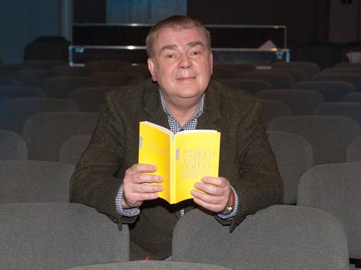 Max Goldt sitzt mit einem gelben Buch in der Hand