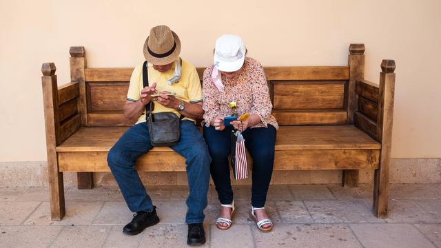 Eine ältere Frau und ein älterer Mann sitzen auf einer Holzbank und blicken auf ihre Smartphones