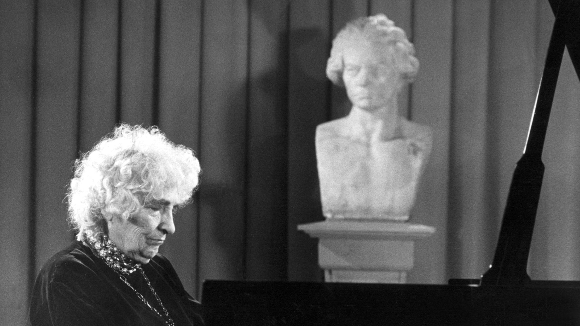Ein schwarz-weiß Foto der Pianistin Elly Ney, zeigt sie am Klavier sitztend und spielend, während ihr eine Büste von Beethoven über die Schulter zu schauen scheint.
