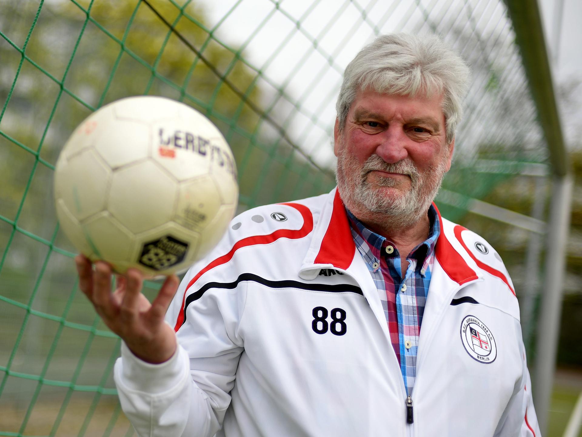 Ein sportlicher älterer Herr mit weißem Haar und Vollbart trägt einen Trainingsanzug mit dem Emblem des Fußballvereins BFC Germania 1888 und hält einen Ball in der rechten Hand.
