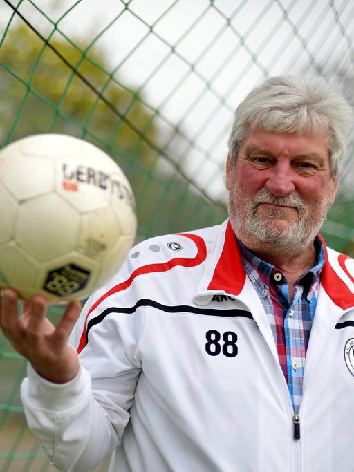 Ein sportlicher älterer Herr mit weißem Haar und Vollbart trägt einen Trainingsanzug mit dem Emblem des Fußballvereins BFC Germania 1888 und hält einen Ball in der rechten Hand.