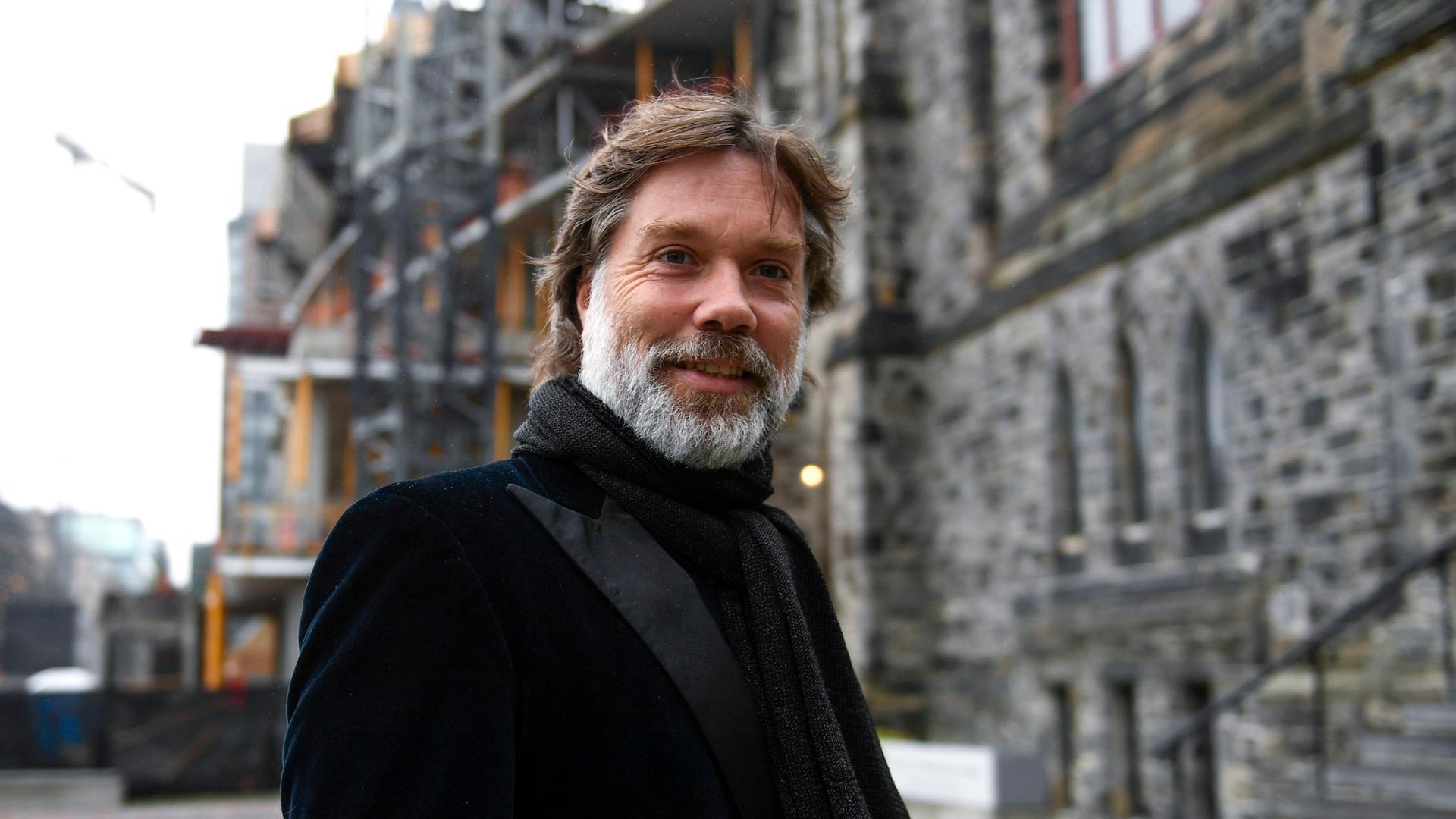 Ein Mann mit braunen Haaren und grauem Vollbart blickt freundlich in die Kamera. Er steht vor einem großen Gebäude, das nur unscharf zu erkennen ist. Er trägt eine schwarze Anzugjacke und einen schwarzen Schal. Ein Mann mit braunen Haaren und grauem Vollbart blickt freundlich in die Kamera. Er steht vor einem großen Gebäude, das nur unscharf zu erkennen ist. Er trägt eine schwarze Anzugjacke und einen schwarzen Schal.
