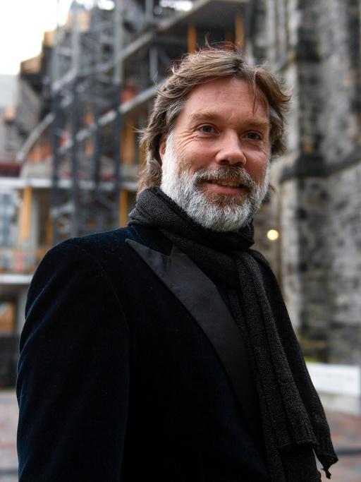 Ein Mann mit braunen Haaren und grauem Vollbart blickt freundlich in die Kamera. Er steht vor einem großen Gebäude, das nur unscharf zu erkennen ist. Er trägt eine schwarze Anzugjacke und einen schwarzen Schal.