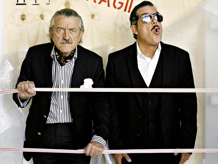 Dieter Meier und Boris Blank von der Band Yello posieren in schwarzen Anzügen vor einer Holzwand mit der Aufschrift "Fragil".