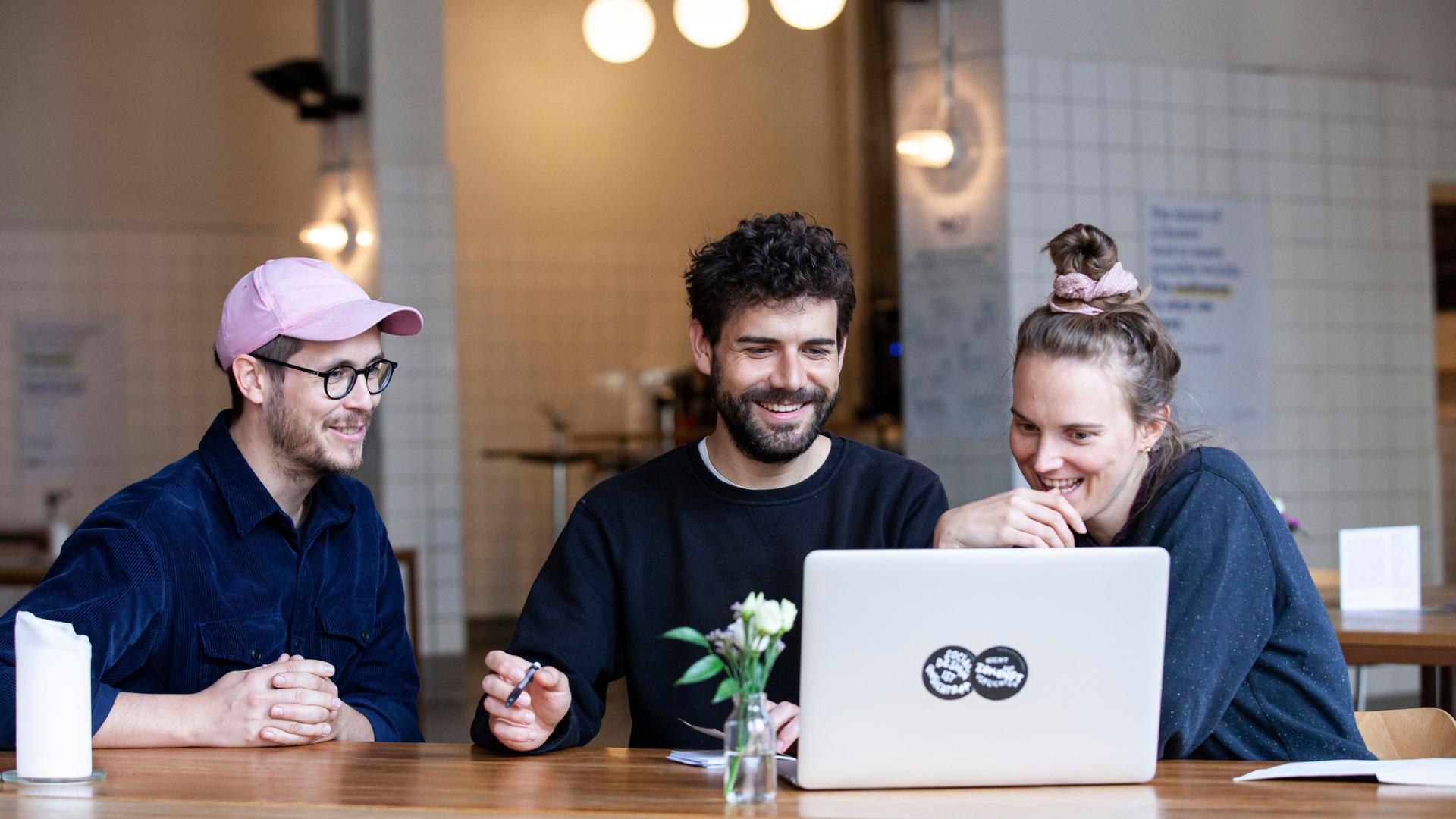 An einem Tisch in einem großen halb-gefliesten Raum sitzen drei Menschen und schauen auf einen Laptop. An einem Tisch in einem großen halb-gefliesten Raum sitzen drei Menschen und schauen auf einen Laptop.