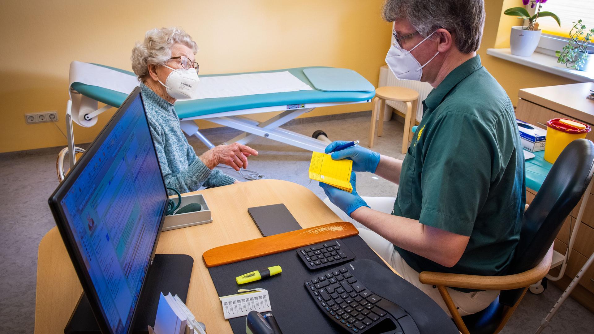 In seiner Hausarztpraxis bespricht Stefan Zutz mit der 95-Jährigen Patientin Ingeborg Ketelhohn die Corona-Impfung, neben ihm auf dem Schreibtisch steht sein PC der alle Patientendaten beinhaltet.