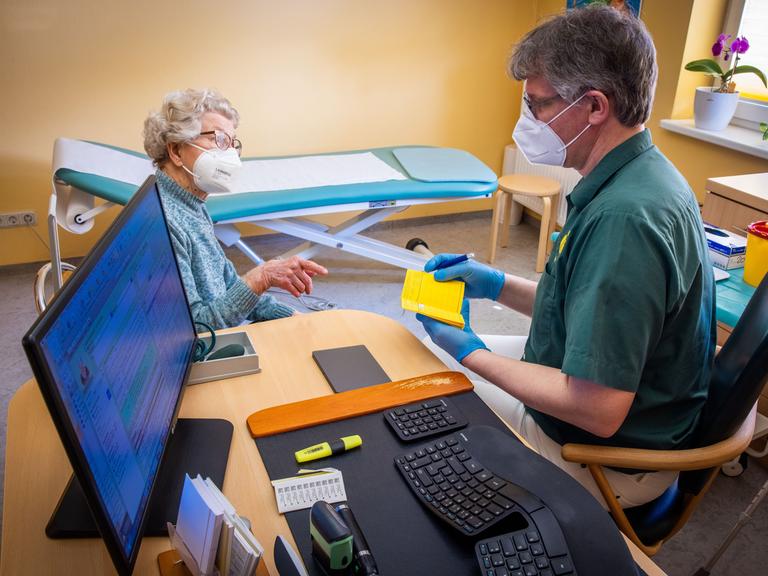 In seiner Hausarztpraxis bespricht Stefan Zutz mit der 95-Jährigen Patientin Ingeborg Ketelhohn die Corona-Impfung, neben ihm auf dem Schreibtisch steht sein PC der alle Patientendaten beinhaltet.