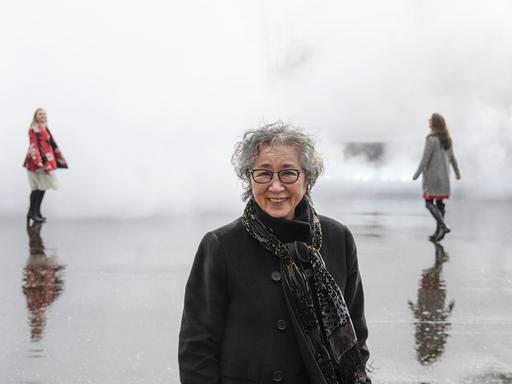 Eine ältere Japanerin mit Brille steht vor eine Nebelwand. Hinter ihr sind die Umrisse zweier Frauen zu erahnen.