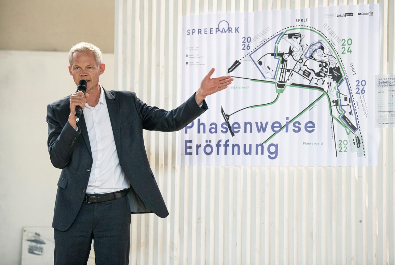 Christoph Schmidt steht vor dem Plan des neuen Spreepark im Plänterwald. Er hat kurze, graue Haare, trägt eine dunklen Anzug und ein weißes Hemd. Im Hintergrund hängt ein Geländeplan des Areals im Osten Berlins.