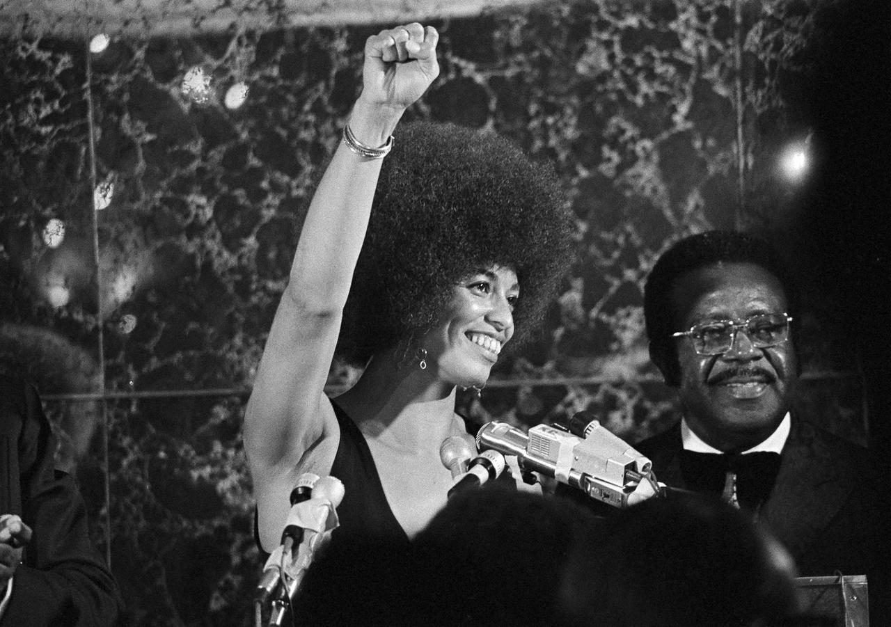Schwarz-weiß-Aufnahme von 1972: Angela Davis steht neben einem afroamerikanischen Mann auf einer Bühne und reckt die Faust zum Gruß der Black-Power-Bewegung.