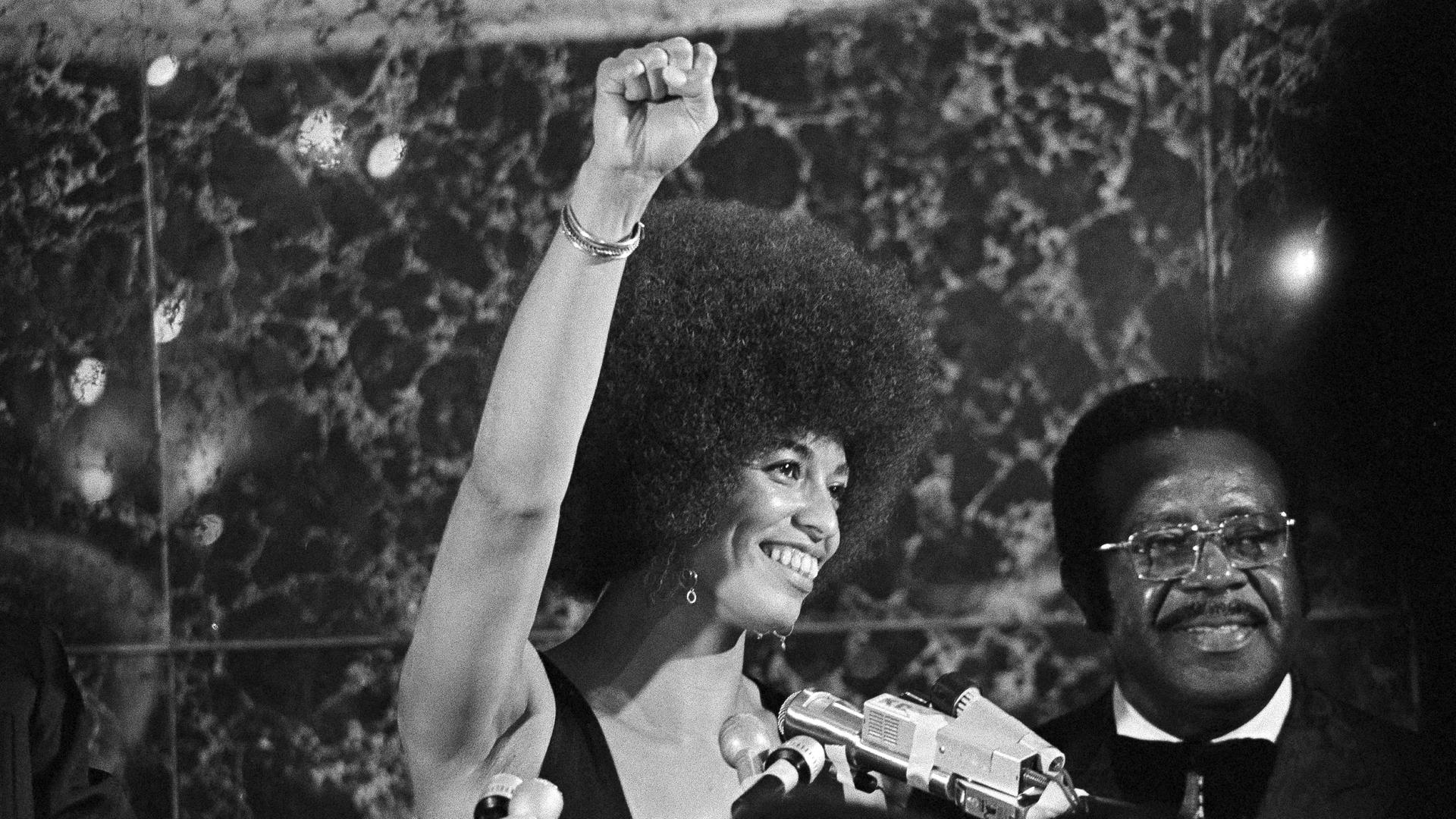 Schwarz-weiß-Aufnahme von 1972: Angela Davis steht neben einem afroamerikanischen Mann auf einer Bühne und reckt die Faust zum Gruß der Black-Power-Bewegung. Schwarz-weiß-Aufnahme von 1972: Angela Davis steht neben einem afroamerikanischen Mann auf einer Bühne und reckt die Faust zum Gruß der Black-Power-Bewegung.