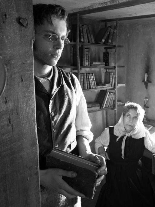 Schwarzweiß-Aufnahme eines Bauernzimmers des 19. Jahrhunderts: Ein junger Mann steht an der Tür, durch die wir eine ältere Frau am Tisch sitzen sehen. Der Gesamteindruck des Bildes ist beengt.