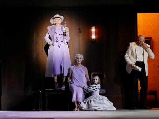 Bei der Inszenierung der Staatsoper Berlin steht eine Frau steht auf einem Podest, auf dem eine ältere Dame sitzt und vor dem ein junges Mädchen kniet. Die drei sollen ein und dieselbe Person in unterschiedlichem Alter darstellen. Links und rechts von ihr stehen Männer in offenen Türrahmen.