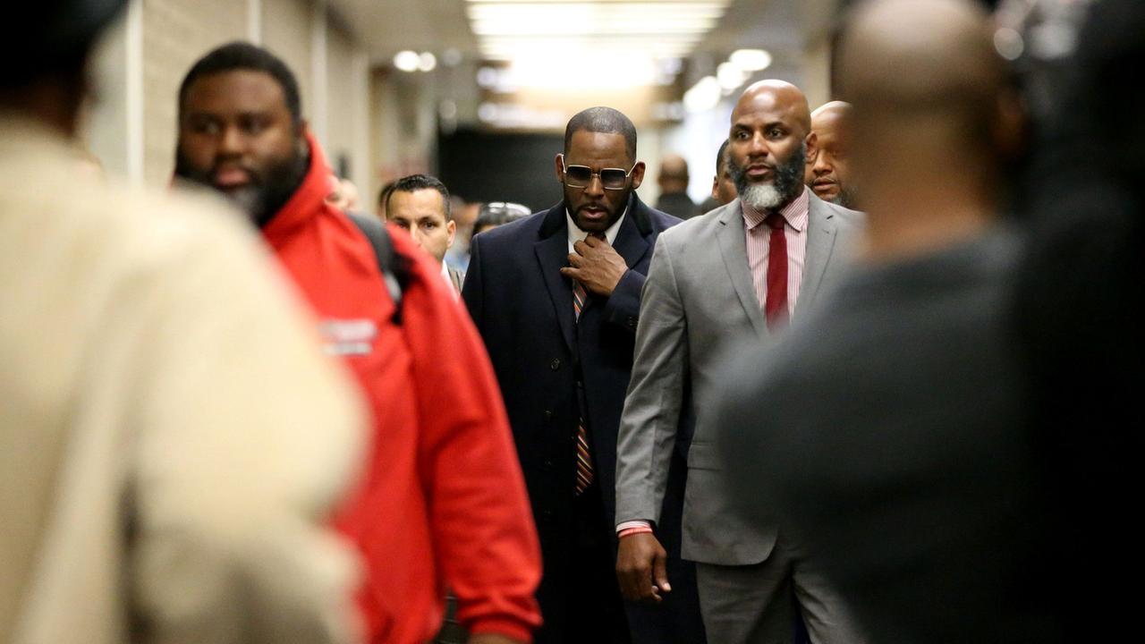 R. Kelly, umgeben von mehreren Personen. Er trägt eine Sonnenbrille und einen dunkelblauen Mantel. Unscharf stehen im Vordergrund zwei Personen mit dem Rücken zur Kamera. R. Kelly, umgeben von mehreren Personen. Er trägt eine Sonnenbrille und einen dunkelblauen Mantel. Unscharf stehen im Vordergrund zwei Personen mit dem Rücken zur Kamera.