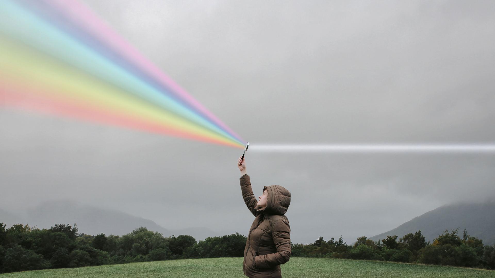 Eine Person steht auf einer Wiese und hält eine Lupe in die Luft in der weißes Licht zum Regenbogenspektrum gebrochen wird. Eine Person steht auf einer Wiese und hält eine Lupe in die Luft in der weißes Licht zum Regenbogenspektrum gebrochen wird.