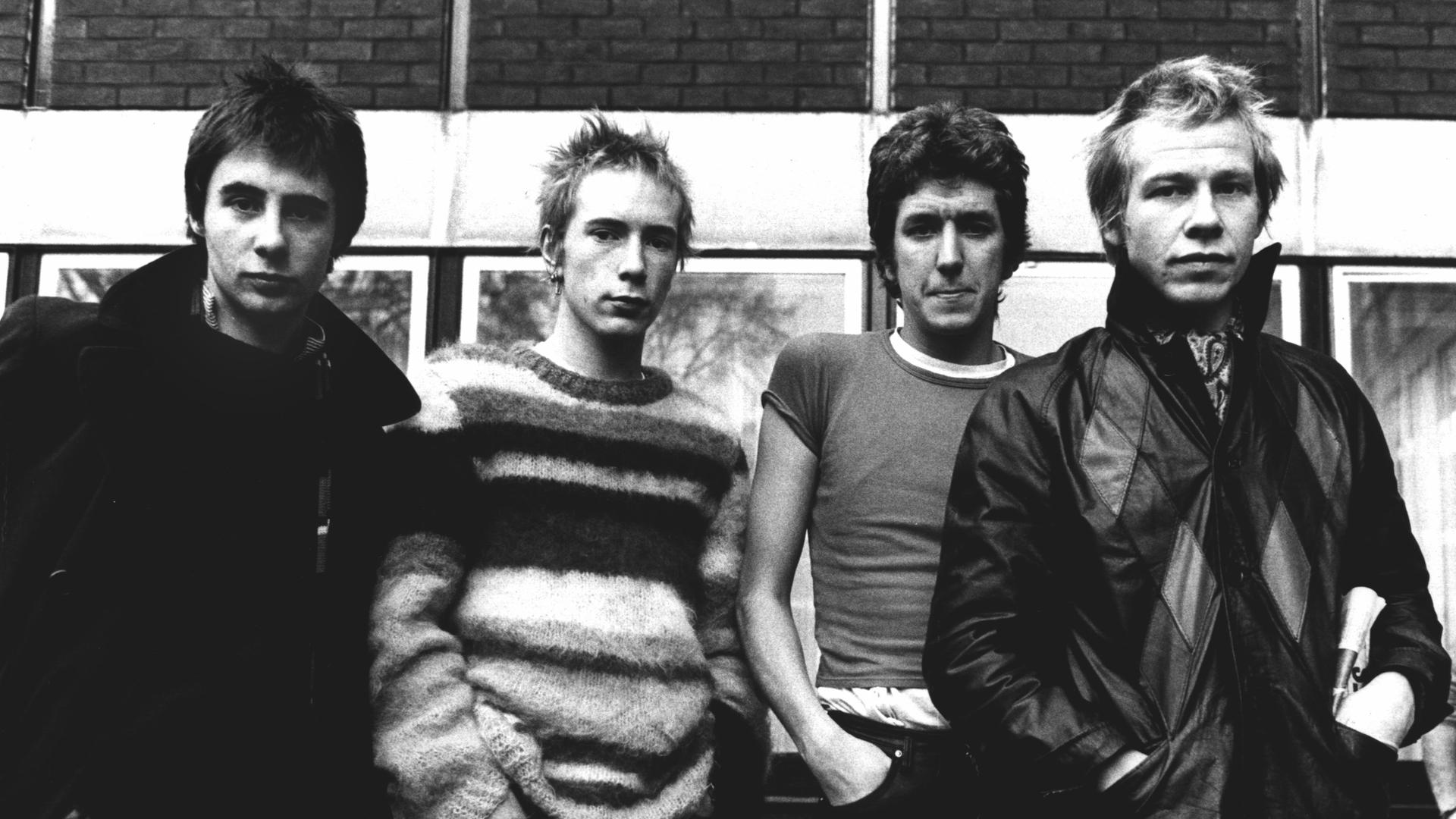 Schwarz-Weiß-Aufnahme der Band Sex Pistols, die nebeneinander vor einem Haus stehen, Hände in den Hosentaschen. Schwarz-Weiß-Aufnahme der Band Sex Pistols, die nebeneinander vor einem Haus stehen, Hände in den Hosentaschen.