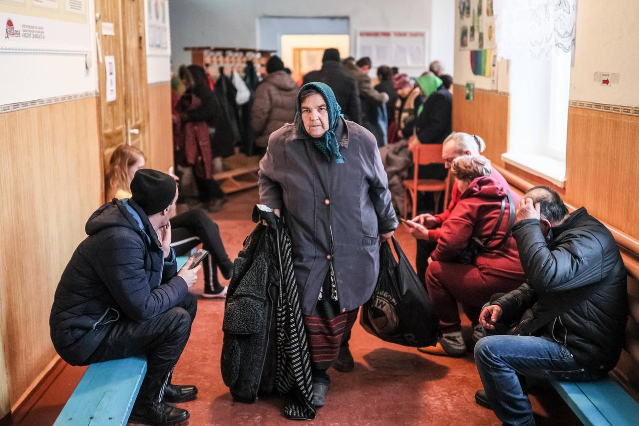 Eine ältere Frau geht durch den Flur eines Schulgebäudes in Mitkovo-Kachkari in der Ukraine. Links und rechts sitzen Menschen auf Bänken.