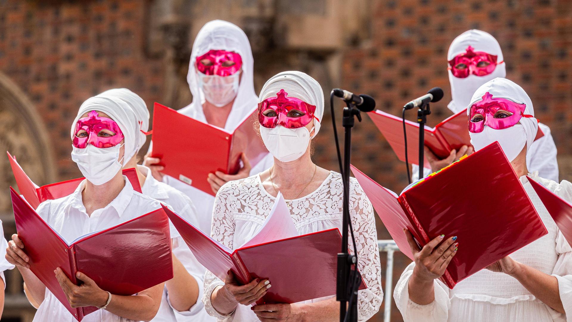 Frauen in weißen Kleidern und weißen, medizinischen Masken und grell roten Augenmasken singen aus großen, roten Mappen. Frauen in weißen Kleidern und weißen, medizinischen Masken und grell roten Augenmasken singen aus großen, roten Mappen.
