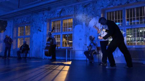 ICI- Musiker spielen in einer dunklen Industriehalle, während durch die vergitterten Fenster Licht von einer Außenbeleuchtung scheint