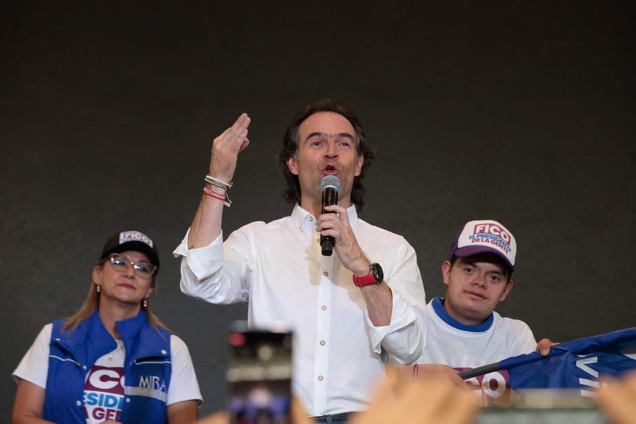 Ein Herr mit etwas längeren Haaren und weißem Hemd gestikuliert zwischen zwei Anhängern mit der Hand und spricht in ein Mikrofon.