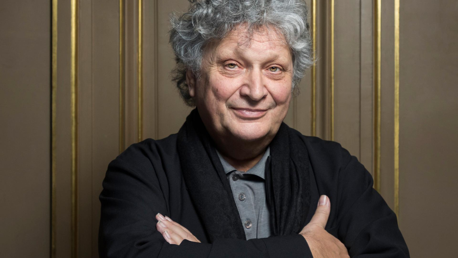 Dirigent René Jacobs steht vor einer edlen Holzvertäfelung mit Teilvergoldungen und hat die Arme verschränkt, während er mit grauen Locken in die Kamera lächelt. Dirigent René Jacobs steht vor einer edlen Holzvertäfelung mit Teilvergoldungen und hat die Arme verschränkt, während er mit grauen Locken in die Kamera lächelt.