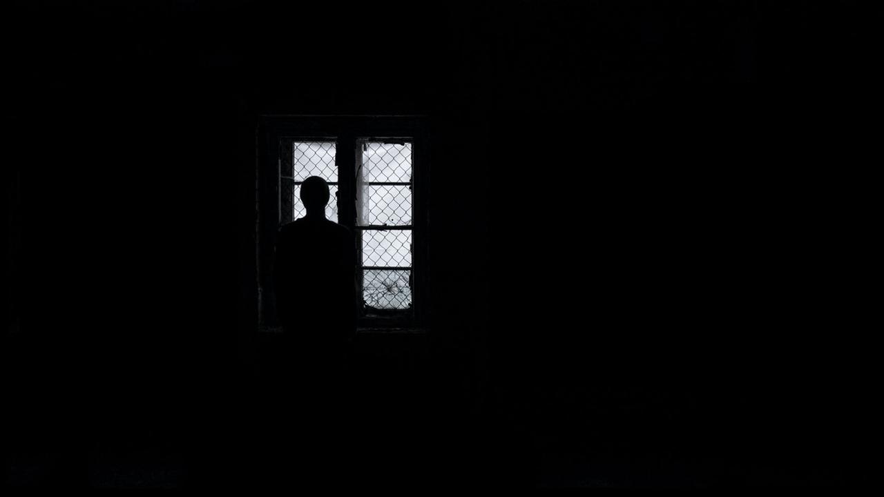 Eine Frau steht als Silhouette im Dunkeln vor vergitterten Fenstern und schaut hinaus Eine Frau steht als Silhouette im Dunkeln vor vergitterten Fenstern und schaut hinaus