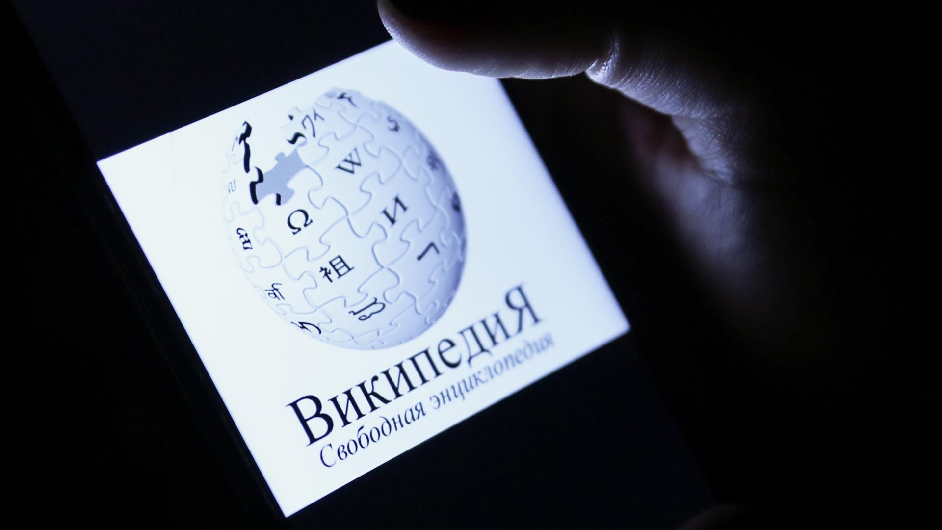 Auf einem Smartphone-Display ist das Logo der Online-Enzyklopädie Wikipedia, darunter der Name der Seite in kyrillischen Buchstaben und in russischer Sprache.