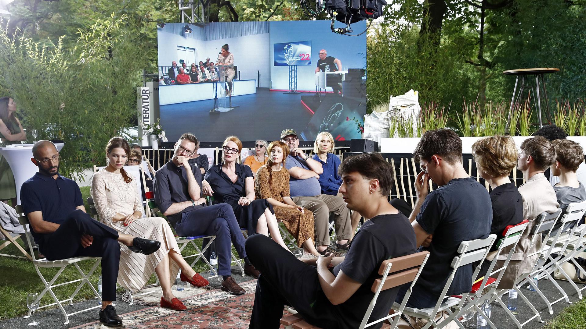 Auf Stühlen sitzen die Teilnehmerinnen und Teilnehmer am Wettbewerb zum Bachmannpreis 2022 am Eröffnungsabend einander gegenüber. Im Hintergrund ist auf einer Leinwand das Innere des ORF-Theaters im Funkhaus Klagenfurt zu sehen, wo die Jury sitzt. Auf Stühlen sitzen die Teilnehmerinnen und Teilnehmer am Wettbewerb zum Bachmannpreis 2022 am Eröffnungsabend einander gegenüber. Im Hintergrund ist auf einer Leinwand das Innere des ORF-Theaters im Funkhaus Klagenfurt zu sehen, wo die Jury sitzt.