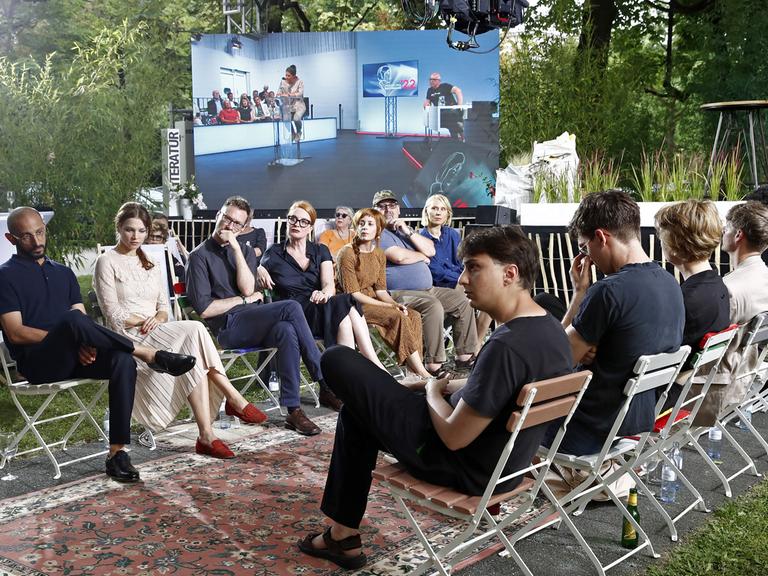 Auf Stühlen sitzen die Teilnehmerinnen und Teilnehmer am Wettbewerb zum Bachmannpreis 2022 am Eröffnungsabend einander gegenüber. Im Hintergrund ist auf einer Leinwand das Innere des ORF-Theaters im Funkhaus Klagenfurt zu sehen, wo die Jury sitzt.