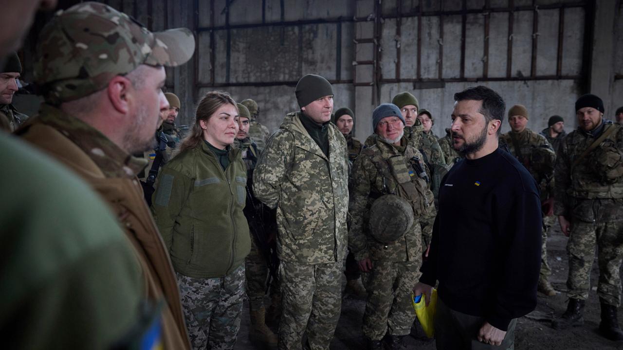 Selenskyj steht vor mehreren Soldaten. Er trägt einen schwarzen Pullover und eine ukrainische Flagge.