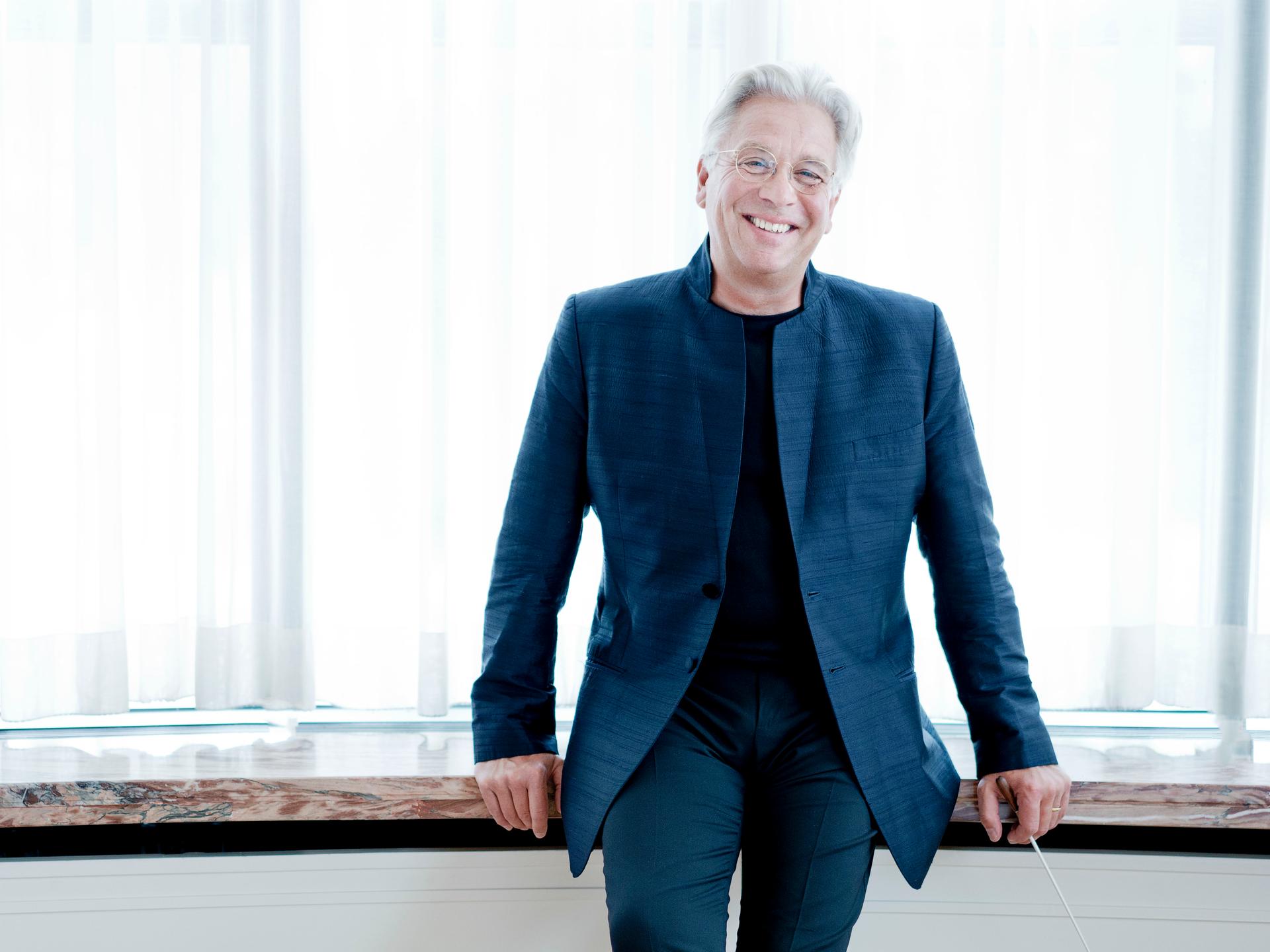 Håkan Hardenberger steht in einem dunkelblauen Anzug vor einem großen Holztisch, an den er sich lachend lehnt. Håkan Hardenberger steht in einem dunkelblauen Anzug vor einem großen Holztisch, an den er sich lachend lehnt.