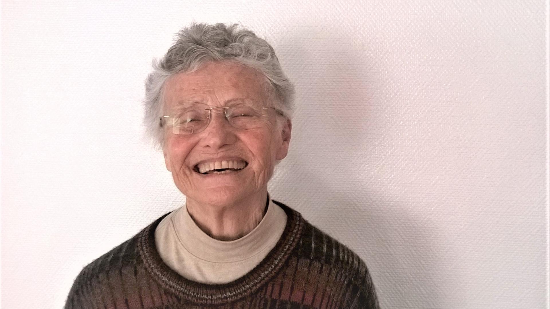 Annea Lockwood ist eine ältere Dame mit kurzen Haaren. Sie steht vor einer weißen Wand und lacht in die Kamera Annea Lockwood ist eine ältere Dame mit kurzen Haaren. Sie steht vor einer weißen Wand und lacht in die Kamera