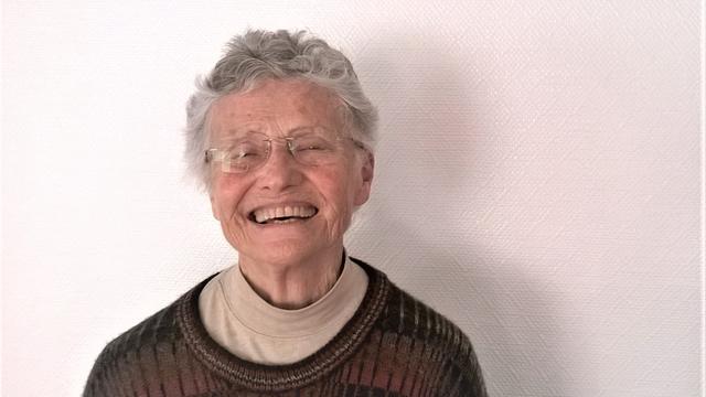 Annea Lockwood ist eine ältere Dame mit kurzen Haaren. Sie steht vor einer weißen Wand und lacht in die Kamera