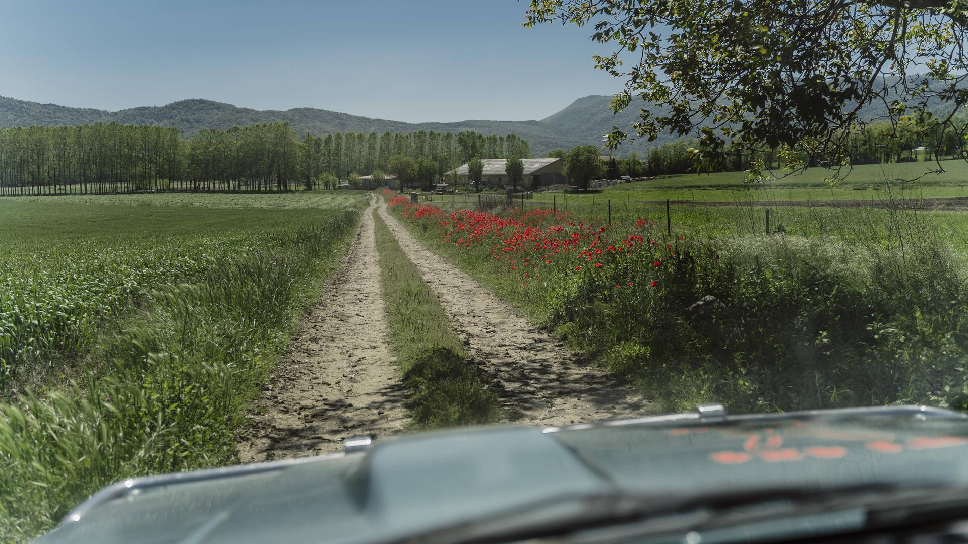 Blick aus einem Auto über die Motorhaube. Das Auto fährt auf einem Feldweg. Im Hintergrund ist ein Bauernhof zu sehen und Berge. Blick aus einem Auto über die Motorhaube. Das Auto fährt auf einem Feldweg. Im Hintergrund ist ein Bauernhof zu sehen und Berge.
