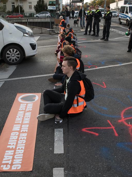 Junge Menschen in orangenen Warnwesten sitzen in einer Reihe auf einer Straße und blockieren den Autoverkehr.