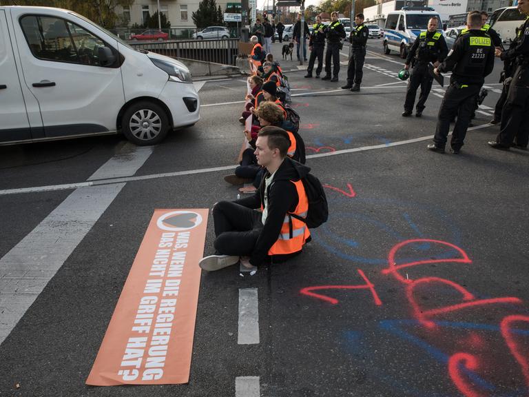 Junge Menschen in orangenen Warnwesten sitzen in einer Reihe auf einer Straße und blockieren den Autoverkehr.