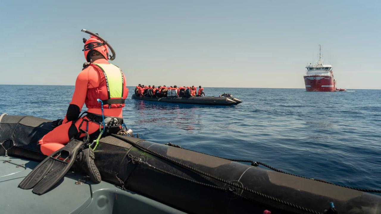 Ein Mann in einem orangenen Taucheranzug sitzt auf einem Schlauchboot und beobachtet ein Boot mit Geflüchteten, das im Meer treibt. Im Hintergrund ist ein Schiff. Das Bild ist vor der Küste der italienischen Insel Lampedusa aufgenommen.