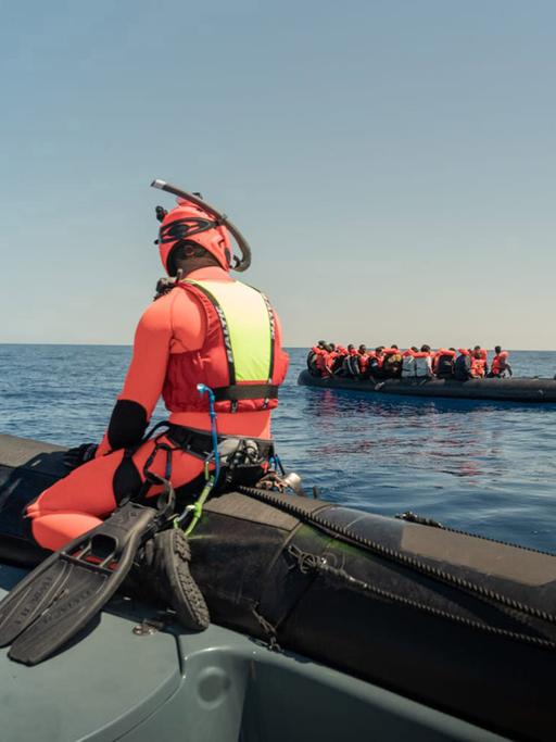 Ein Mann in einem orangenen Taucheranzug sitzt auf einem Schlauchboot und beobachtet ein Boot mit Geflüchteten, das im Meer treibt. Im Hintergrund ist ein Schiff. Das Bild ist vor der Küste der italienischen Insel Lampedusa aufgenommen.
