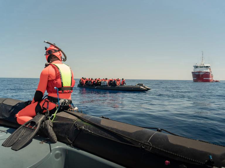 Ein Mann in einem orangenen Taucheranzug sitzt auf einem Schlauchboot und beobachtet ein Boot mit Geflüchteten, das im Meer treibt. Im Hintergrund ist ein Schiff. Das Bild ist vor der Küste der italienischen Insel Lampedusa aufgenommen.