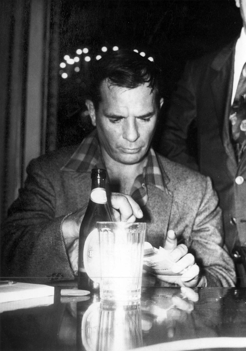 Porträt von Jack Kerouac, der an einem Tisch sitzt und eine Zigarette aus der Schachtel zieht, vor sich eine Bierflasche und ein Glas.