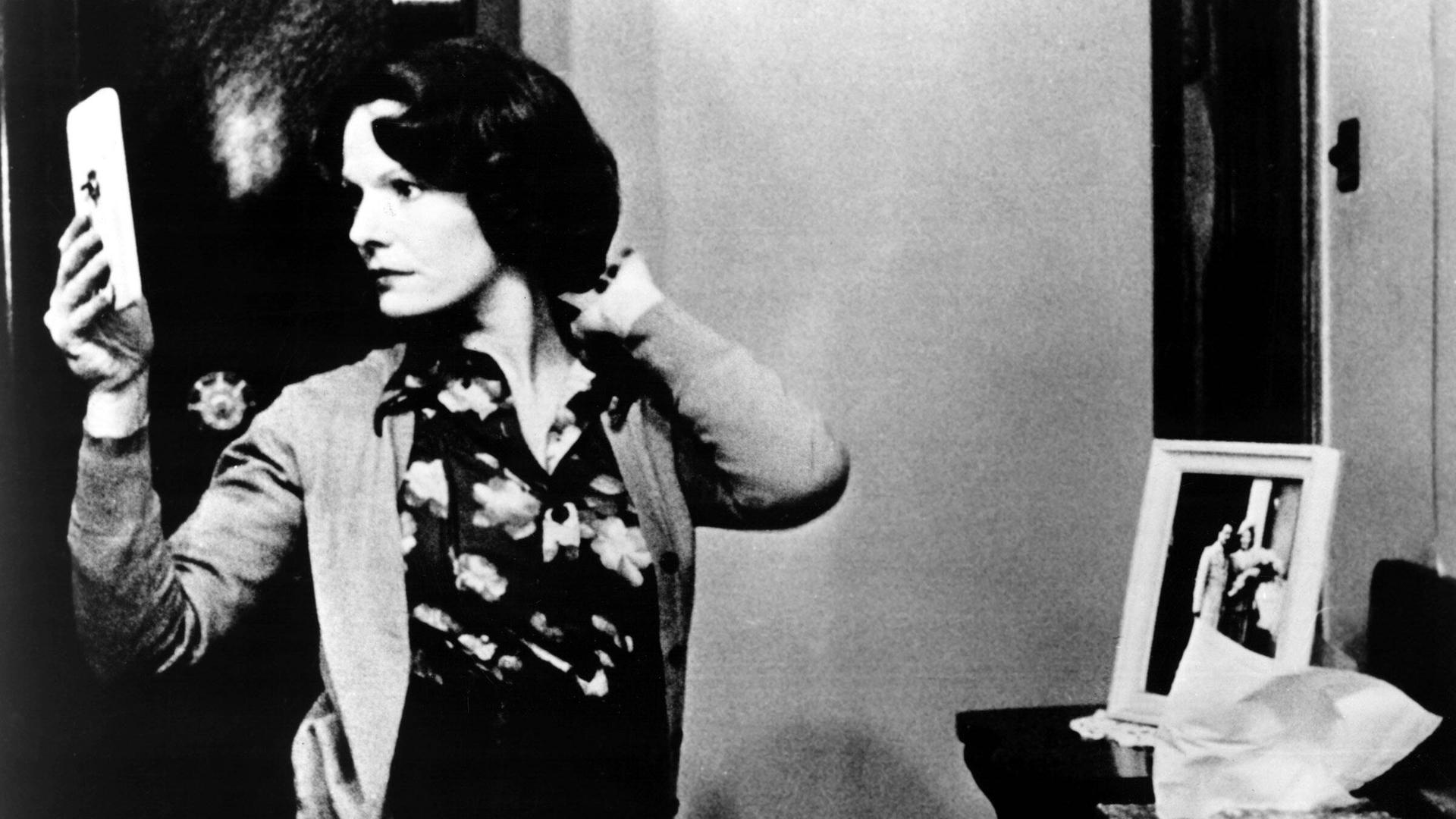 Die Szene aus dem Film "Jeanne Dielman" zeigt in Schwarz-Weiß eine Frau in einem Raum, die sich in einem Handspiegel betrachtet.