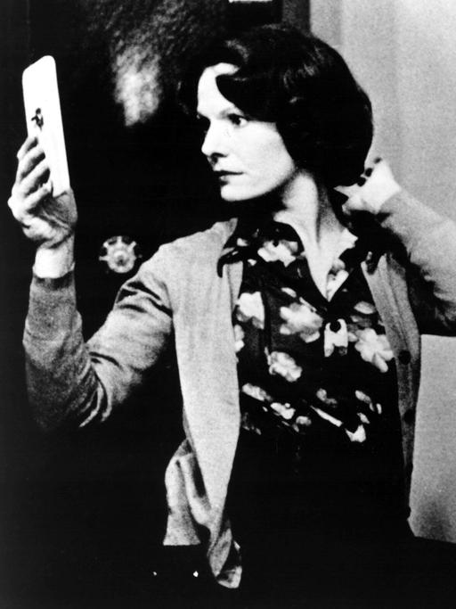 Die Szene aus dem Film "Jeanne Dielman" zeigt in Schwarz-Weiß eine Frau in einem Raum, die sich in einem Handspiegel betrachtet.