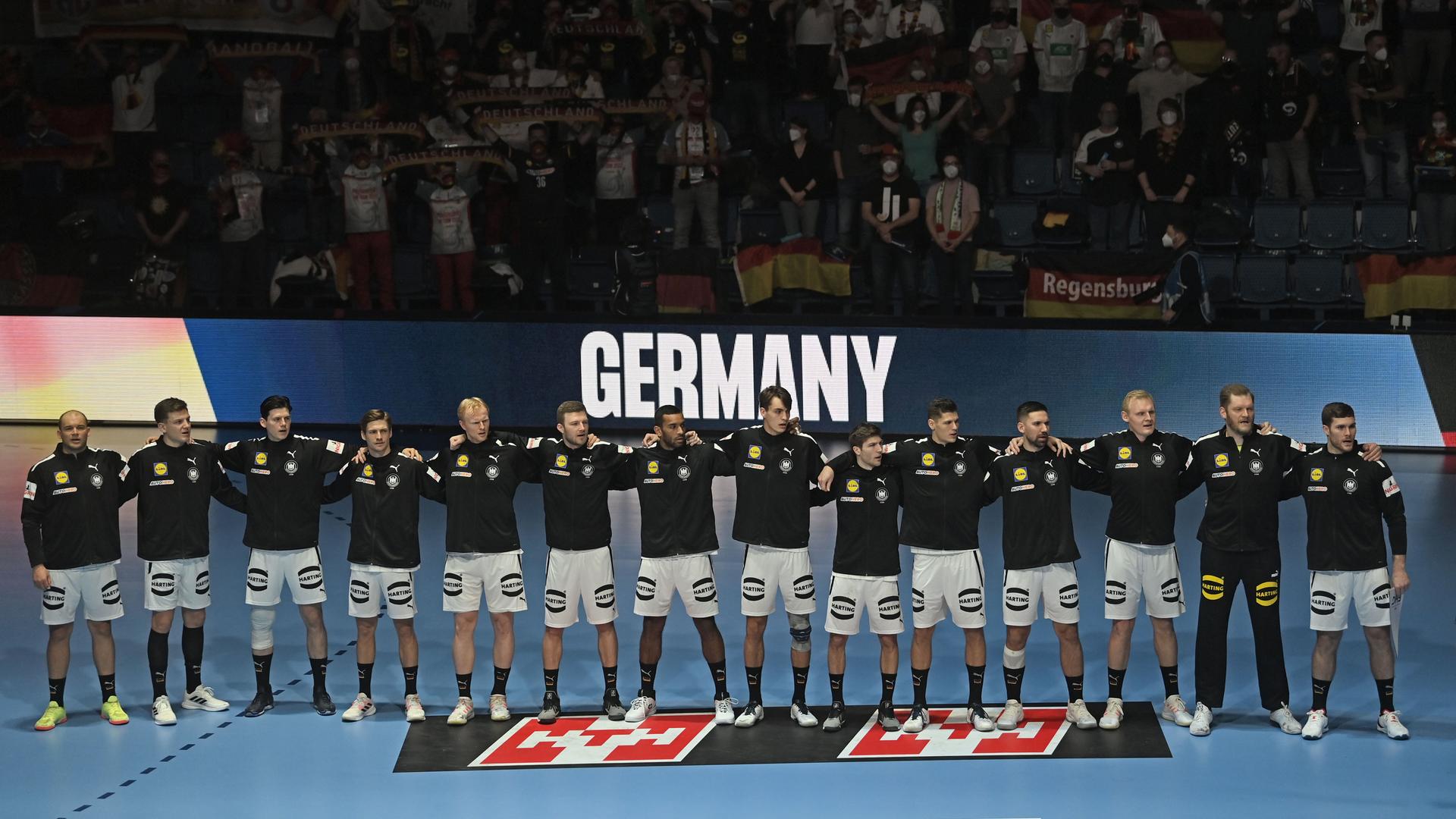 Die Spieler in schwarzen Trainingsjacken stehen nebeneinander und Arm in Arm auf dem Platz. Hinter ihnen der Schriftzug "Germany" und Zuschauer im Dunkeln auf den Tribünen. Manche davon mit Deutschland-Fahnen. Die Spieler in schwarzen Trainingsjacken stehen nebeneinander und Arm in Arm auf dem Platz. Hinter ihnen der Schriftzug "Germany" und Zuschauer im Dunkeln auf den Tribünen. Manche davon mit Deutschland-Fahnen.
