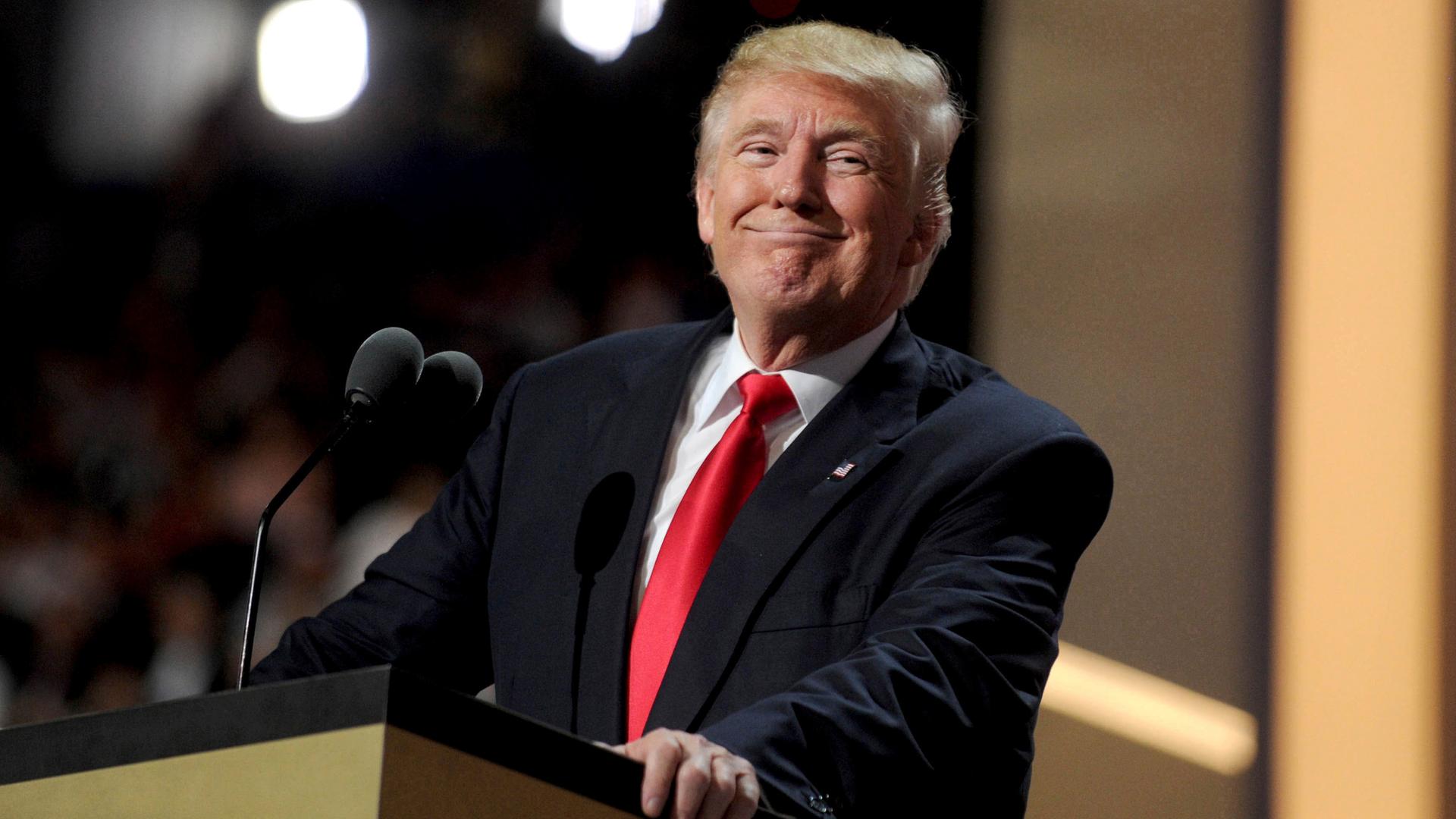 Donald Trump steht hinter einem Rednerpult und lächelt breit.