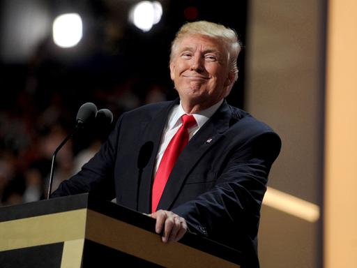 Donald Trump steht hinter einem Rednerpult und lächelt breit.