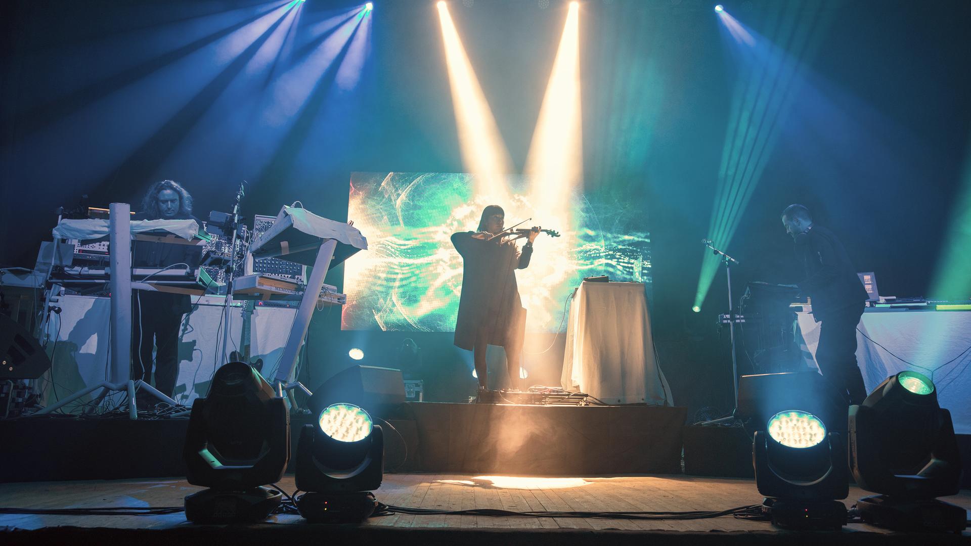 Die drei Mitglieder von Tangerine Dream performen auf einer grün-blau beleuchteten Bühne. Die drei Mitglieder von Tangerine Dream performen auf einer grün-blau beleuchteten Bühne.