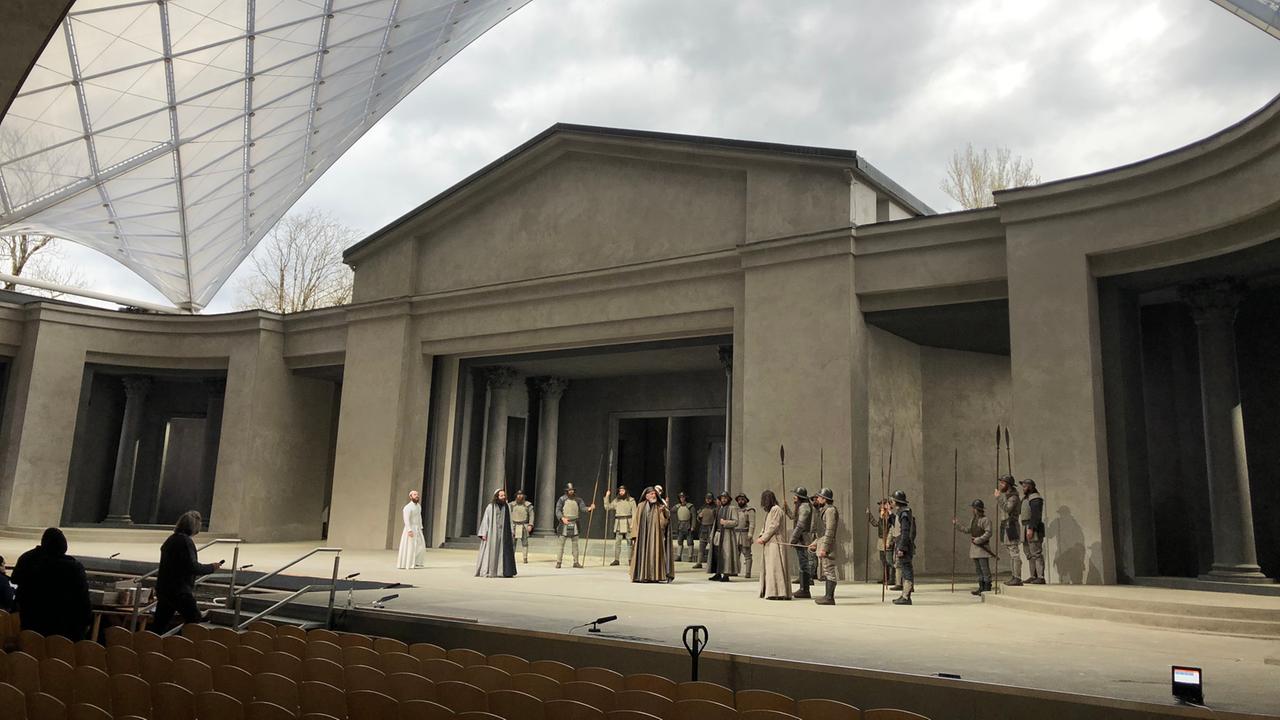 Auf der Bühne in Oberammergau stehen Dutzende Schauspieler. Das Dach für das Publikum wölbt sich links im Bild, über der Bühne ist der Himmel zu sehen, ganz im Vordergrund Stühle. Auf der Bühne in Oberammergau stehen Dutzende Schauspieler. Das Dach für das Publikum wölbt sich links im Bild, über der Bühne ist der Himmel zu sehen, ganz im Vordergrund Stühle.
