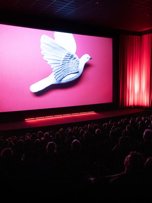Eröffnungszeremonie des Filmfestivals DOK Leipzig. Zu sehen ist ein voller Kinosaal mit einer großen Taube auf der Leinwand.