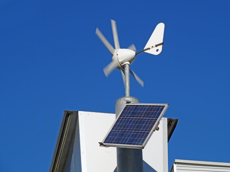 Weiße Miniwindräder und Solar Panels auf einem Häuserdach vor blauem Himmel.